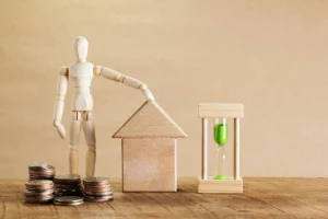 Wooden house model with stacked coins and hourglass representing cost considerations and budget planning for building with hempcrete in India.
